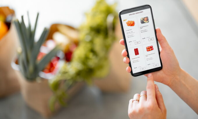Holding phone with running e-shop application and bags full of fresh food on the background. Shopping groceries online concept